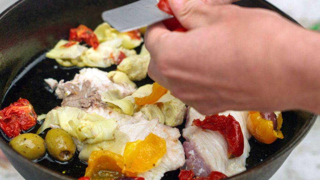 Close up of Chef Corso cutting a tomato into a pan of fried fish and vegetables.
