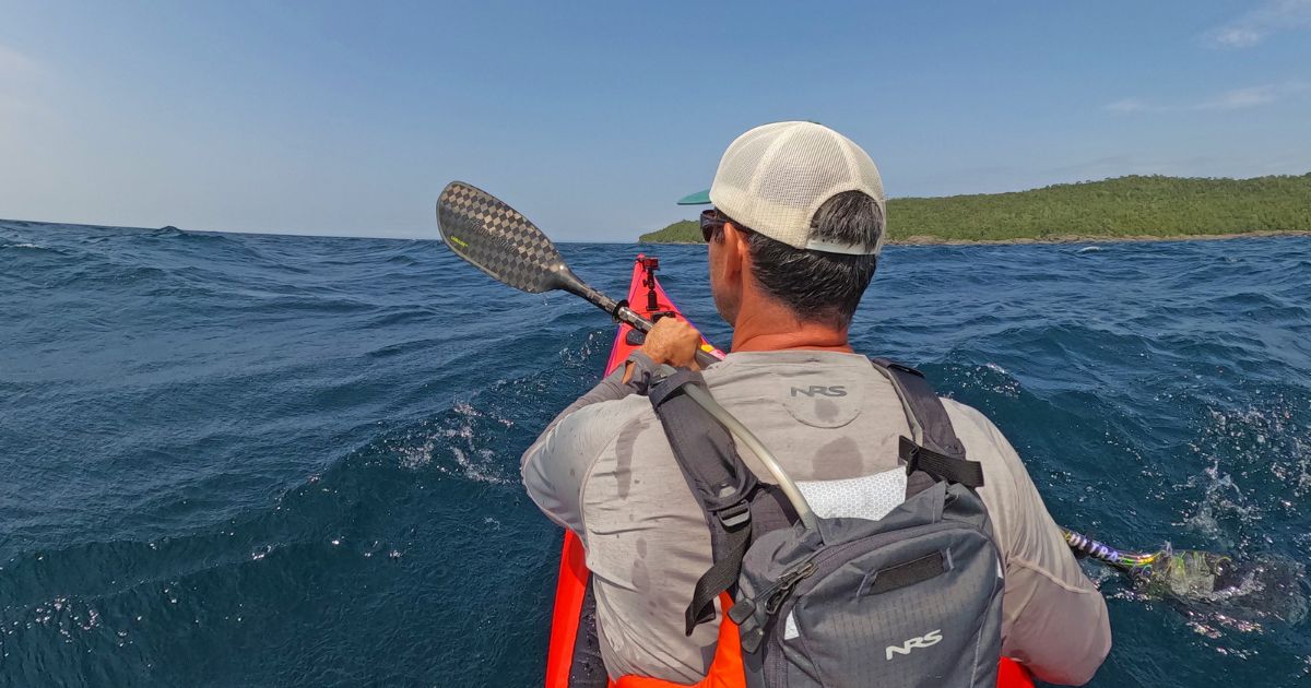 Ken Whiting paddling a kayak across the rough, but clear waters of Lake Superior, surrounded by dense forest.