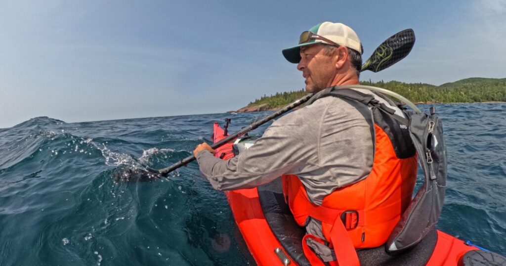 Ken Whiting paddling a kayak across the rough, but clear waters of Lake Superior, surrounded by dense forest.