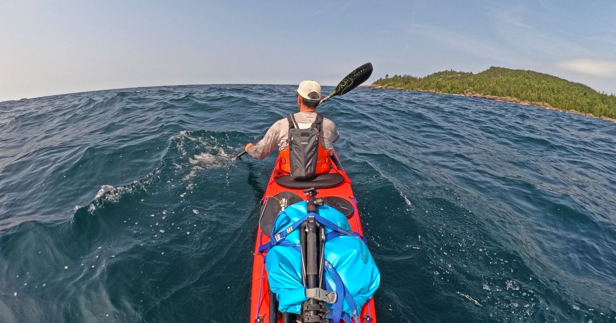 Ken Whiting paddling a kayak across the rough, but clear waters of Lake Superior, surrounded by dense forest.