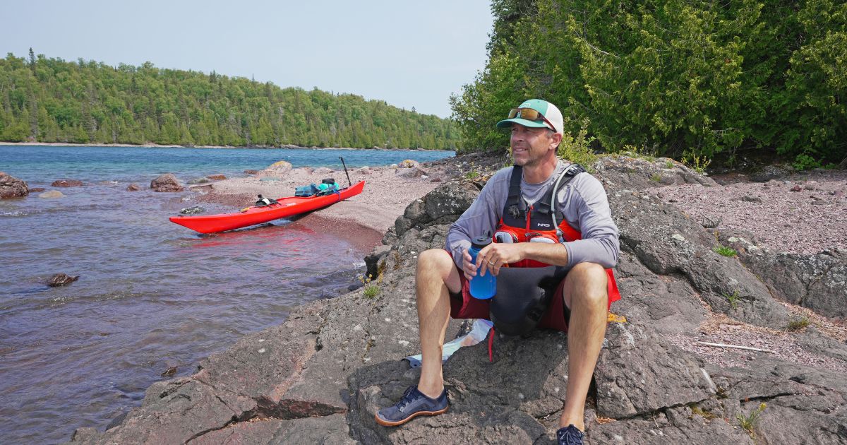 Ken Whiting paddling a kayak across the calm, clear waters of Lake Superior, surrounded by rugged cliffs and dense forest.