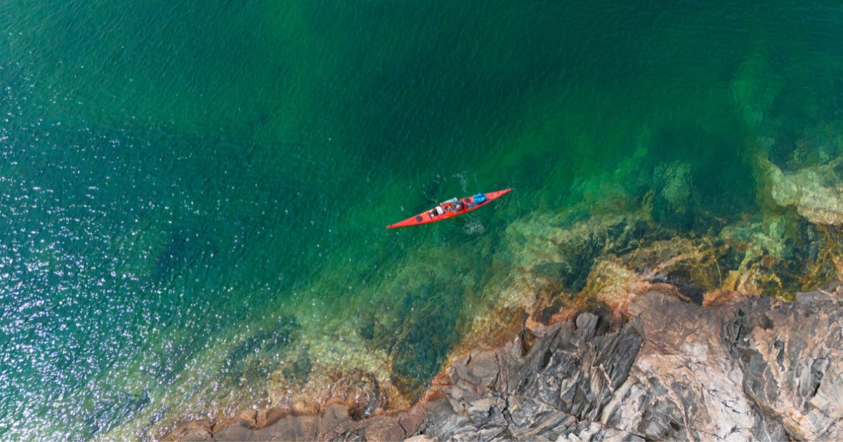 Ken Whiting paddling a kayak across the calm, clear waters of Lake Superior, surrounded by rugged rocks and water.