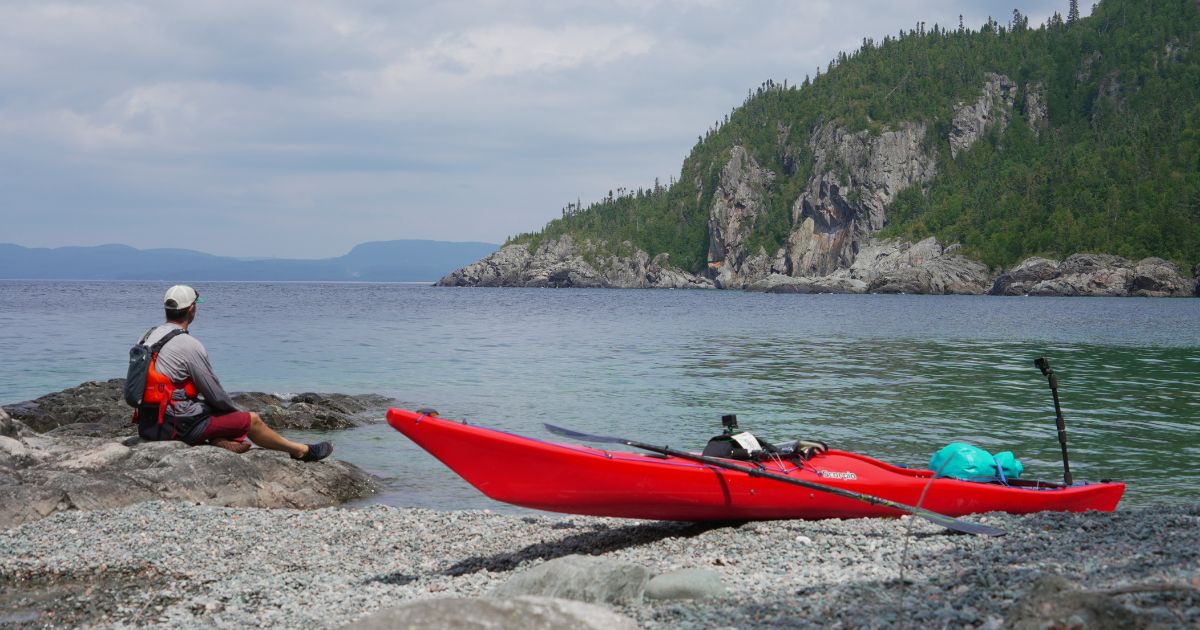 Ken Whiting sitting on the shoreline with a kayak silhouetted in the foreground, emphasizing the tranquility of solo camping.